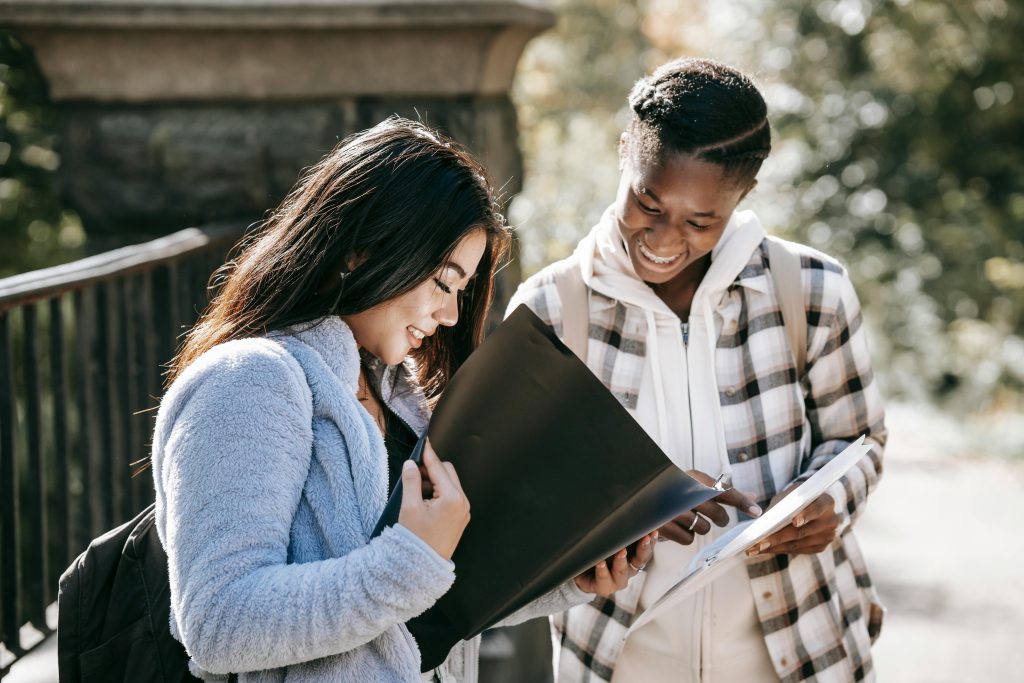 Two diverse students smiling and sharing study materials in a sunny outdoor setting.